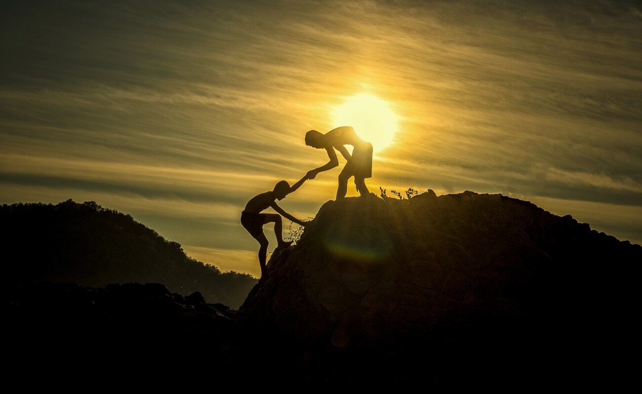 朝日の前で2人の人間が支えあって山を登っている写真